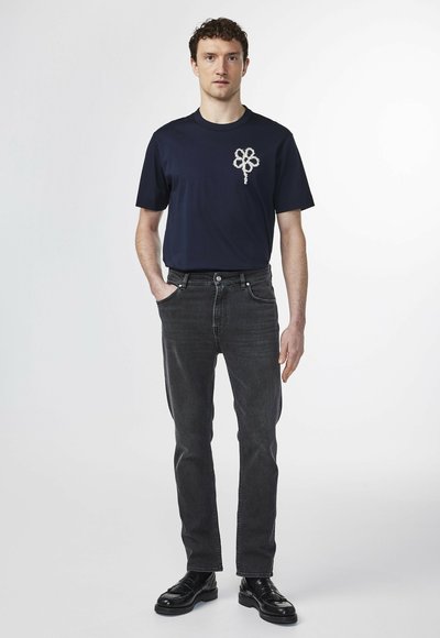Navy t-shirt with a textured flower design on the left chest, paired with dark gray jeans and black dress shoes.