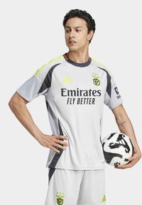 Fodboldtrøje i lys grå med sorte og neongule accenter. Har logo og teksten "Emirates Fly Better". Spiller holder en sort og hvid fodbold.