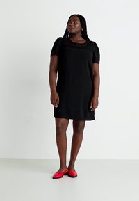 Robe noire à manches courtes avec un empiècement en dentelle texturée, coupe décontractée et longueur de genoux. Associée à des chaussures plates rouges sur un fond uni.