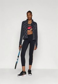 Γυναίκα σε μαύρα αθλητικά ρούχα και λουλουδάτο σουτιέν, κρατάει μια ρακέτα τένις, στέκοντας μπροστά σε απλό φωτεινό φόντο.