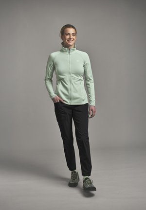 Lächelnde Frau geht, trägt eine hellgrüne Jacke mit Reißverschluss, schwarze Hose und grüne Wanderschuhe vor einem schlichten grauen Hintergrund.