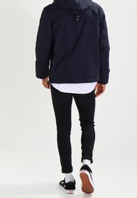 Chaqueta con capucha navy, camisa blanca debajo, pantalones negros ajustados y zapatillas negras con acentos blancos y suelas estampadas. Conjunto casual visto desde atrás.
