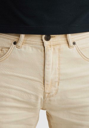 Gros plan sur la taille d'un jean beige avec fermeture par bouton, passants pour ceinture, poches avant, porté avec un haut noir.