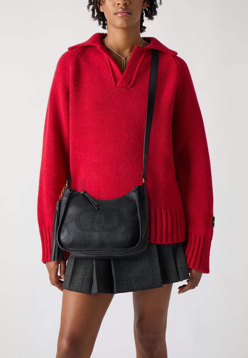 Jersey rojo de punto con escote en V, combinado con un bolso de hombro de cuero negro texturizado que presenta una correa y un detalle de fleco. Falda de cuadros visible.
