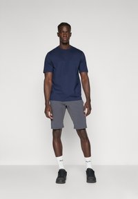 Marineblaues T-Shirt mit kurzen Ärmeln und geradem Saum sowie dezentem Branding; kombiniert mit hellgrauen Shorts und schwarzen Turnschuhen, weißen Knöchelsocken.