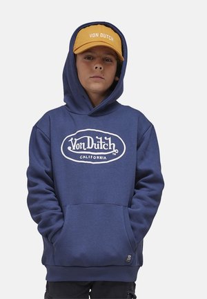 Garçon portant un sweat à capuche Von Dutch bleu marine et une casquette Von Dutch jaune moutarde, debout les mains dans les poches du sweat à capuche, expression neutre.