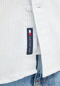 Camicia striped bianca e azzurro chiaro con chiusura a bottoni, dotata di un'etichetta navy con la dicitura "1985 TOMMY JEANS" e accenti rossi e bianchi.