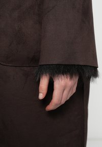 Καφέ σουέτ ύφασμα με μαύρες μανσέτες από ψεύτικη γούνα. Το μανίκι έχει λεία υφή, ενώ η γούνα έχει αφράτη, υφή εμφάνιση.