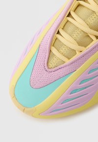 Gros plan sur la zone des orteils d'une basket avec des lacets jaune pastel, une tige en mesh et des superpositions synthétiques violettes, turquoise et jaunes sur fond blanc.