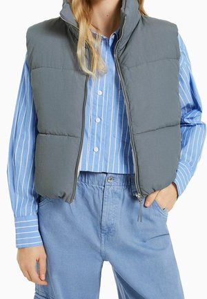 Personne portant un gilet matelassé gris sur une chemise à rayures bleues et blanches boutonnée et un pantalon bleu clair, avec une main dans la poche.