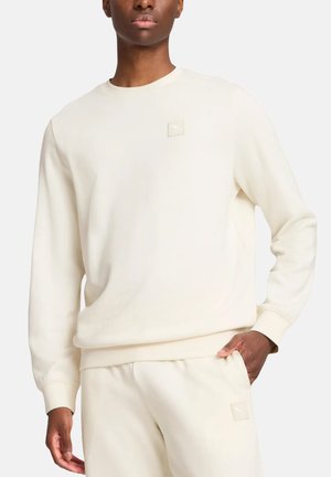 Uomo con felpa color crema e pantaloni della tuta abbinati con sottili patch con logo sul petto e sulla coscia, in piedi con la mano in tasca.