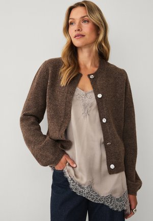 Woman with long blonde hair wearing a brown buttoned cardigan over a beige lace-trimmed top and dark pants, posing with one hand in pocket.
