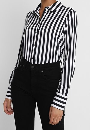 Žena v černobílé košili s vertikálními pruhy zapnuté na knoflíky, zastrčené do vysoké černé kalhoty, pózuje na jednobarevném pozadí.