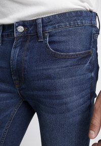 Tummansinisiä farkkuja, joissa on kapea leikkaus. Ominaisuuksiin kuuluvat etutasku, jossa on ompeluyksityiskohtia, metallinappi ja hieman kulunut pinta.