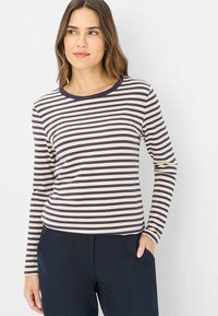 Lange mouw gestreepte shirt in beige en marineblauw, met een ronde halslijn en zachte stof. Draagt een marineblauwe broek, met de handen in de zakken.