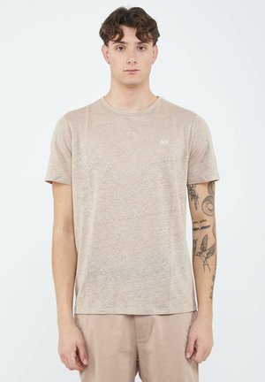 Giovane uomo con capelli ricci che indossa una maglietta beige e pantaloncini, con tatuaggi visibili sul braccio destro, in piedi davanti a uno sfondo bianco uniforme.