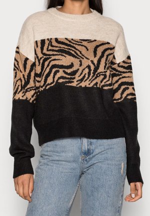 Vrouw draagt een beige, tan en zwarte trui met abstract dierenprint en lichtblauwe gerafelde spijkerbroek, staand tegen een effen achtergrond.