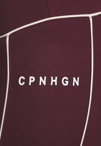 Tessuto bordeaux con piping bianco e lettere bianche "CPNHGN" centrate sul pannello, probabilmente parte di un indumento o di un accessorio.