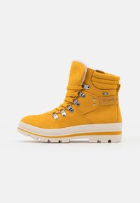 Bottes cheville en daim jaune avec un col rembourré, des œillets métalliques, et des semelles en caoutchouc blanc, arborant un logo latéral et une bande jaune contrastante.