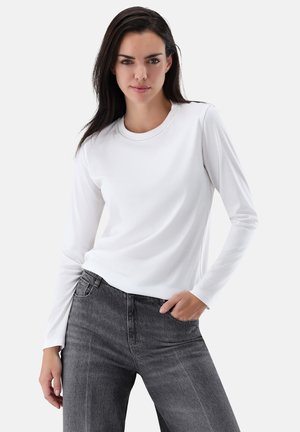 Long-sleeve white top in soft fabric, round neckline, worn with high-waisted gray jeans. Features smooth texture and fitted design.