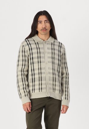Knapp-opp cardigan i lys beige med et teksturert strikkemønster. Har mørkegrønne og svarte striper langs torso og ermer.