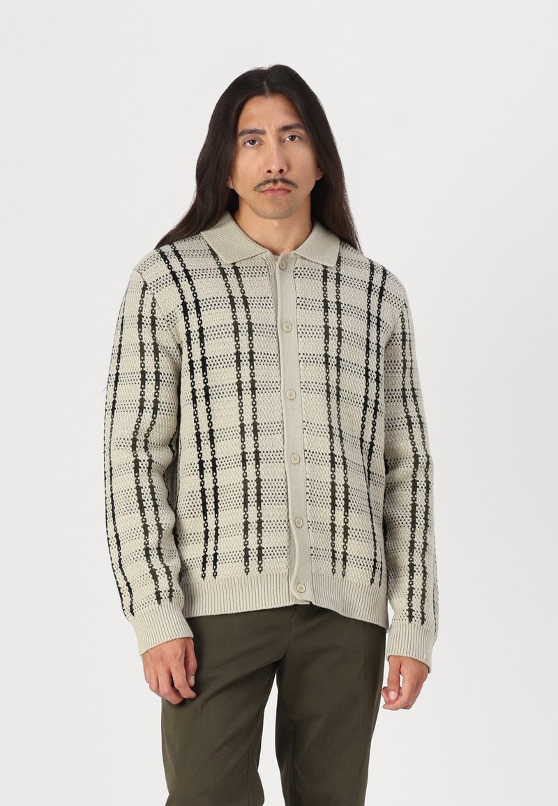 Knapp-opp cardigan i lys beige med et teksturert strikkemønster. Har mørkegrønne og svarte striper langs torso og ermer.