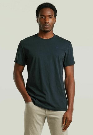 Mann mit kurzen lockigen Haaren, der ein schwarzes T-Shirt und beige Hose trägt, steht mit einer Hand in der Tasche vor einem schlichten Hintergrund.