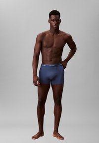 Boxer brevissimi in cotone blu con un'elastico con logo, indossati da un modello maschile su uno sfondo neutro.