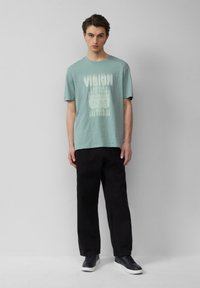 Himmelblaues Baumwoll-T-Shirt mit weißem "VISION"-Grafik und Städtenamen, kombiniert mit schwarzen Hosen und schwarzen Sneakers. Lässige Passform und kurze Ärmel.