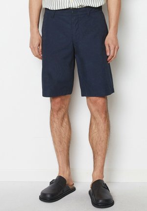 Mand iført mørkeblå knæ-lange shorts, en stribet skjorte stoppet ind i bukserne og sorte slip-on sko, står mod en hvid væg.