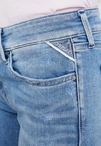 Jeans de mezclilla de color azul claro con una textura desgastada, que cuentan con una etiqueta de logo, diseño de cinco bolsillos y accesorios de metal en la cintura.