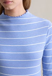 Blaues, geripptes Oberteil mit weißen horizontalen Streifen, das einen hohen Ausschnitt mit einem Rüschensaum aufweist. Eine Nahaufnahme zeigt die Textur und das Muster.