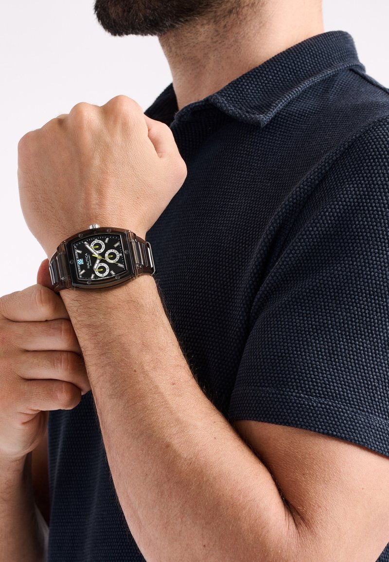Ceas rectangular maro cu un cadran negru, accente albe și galbene, și brățară din metal. Este purtat pe un încheietură, asortat cu un tricou polo texturat de culoare bleumarin.