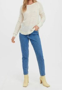 Pull en tricot crème clair avec un motif texturé, associé à un jean en denim bleu et des bottines beiges à chevilles. Le pull présente un col rond et des poignets côtelés.