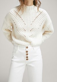 Nainen, joka on pukeutunut valkoiseen, paksuun neulepaitaan, jossa on V-muotoista yksityiskohtaa, yhdistettynä korkeavyötäröisiin valkoisiin housuihin, joissa on viisi puista nappia.