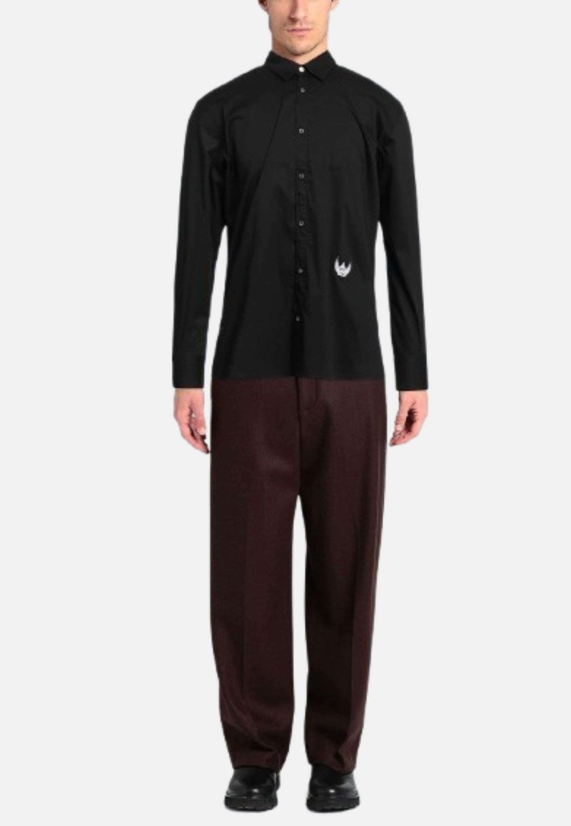 Uomo che indossa una camicia nera con bottoni e un piccolo logo di fiamma bianca, con pantaloni larghi marrone scuro, in piedi su uno sfondo semplice.