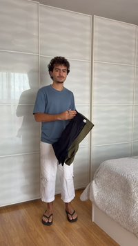 Homme debout dans la chambre près du lit et des portes du placard, tenant deux paires de shorts, portant un t-shirt bleu, un pantalon blanc et des tongs noires.