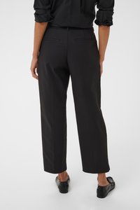 Persona che indossa pantaloni neri a gamba dritta e mocassini neri, in piedi con le braccia rilassate lungo i fianchi, vista da dietro su uno sfondo uniforme.