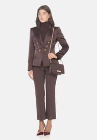 Blazer doppiopetto in satin marrone con bottoni dorati, abbinato a pantaloni fitted coordinati. Accessoriato con una piccola borsa marrone e tacchi bordeaux.