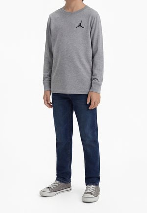 Barn iført en grå langærmet skjorte med et sort logo, mørkeblå jeans og grå snøresneakers, står mod en hvid baggrund.