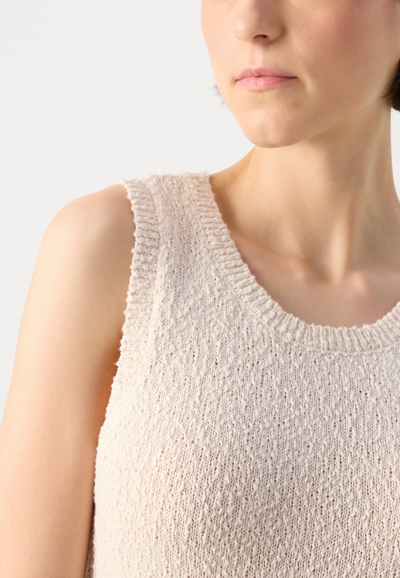 Débardeur en tricot beige clair sans manches avec un col côtelé et un motif texturé. Le tissu semble doux et légèrement tricoté lâche.