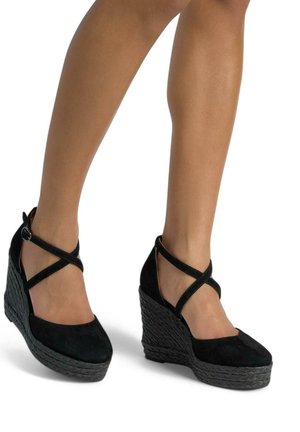 Scarpe con zeppa in suede nero, dotate di cinturini incrociati alla caviglia e una suola a piattaforma tessuta texture. Il design enfatizza l'altezza e la stabilità.