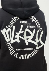 Sudadera negra con el logo de graffiti blanco "DKRY", la frase "Conocido por las calles" y texto gótico circular "Respetador sin miedo" y "Pureza y autenticidad".