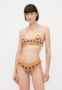 Nertas nėrinių bikinis, turintis trikampį viršų ir žemai įsiuvamus šortus, smėlinės spalvos, su margais languotais raštais ir banguotais kraštais.