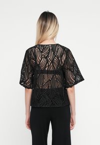 Svart blonderbluse med gjennomsiktige detaljer, med bølgete mønster og korte ermer. Designet inkluderer intrikate teksturer og bølgete aksenter.