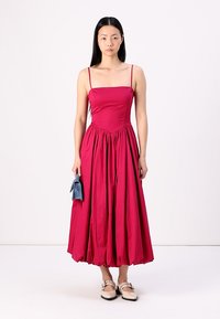 Vestido midi fucsia con tirantes finos, cuerpo ajustado, falda fluida y tejido texturizado. La modelo sostiene un bolso pequeño azul y lleva zapatos planos.