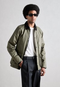 Chaqueta ligera verde oliva con cuello negro, botones en el frente y cierre de cremallera, combinada con una camiseta blanca y pantalones negros.
