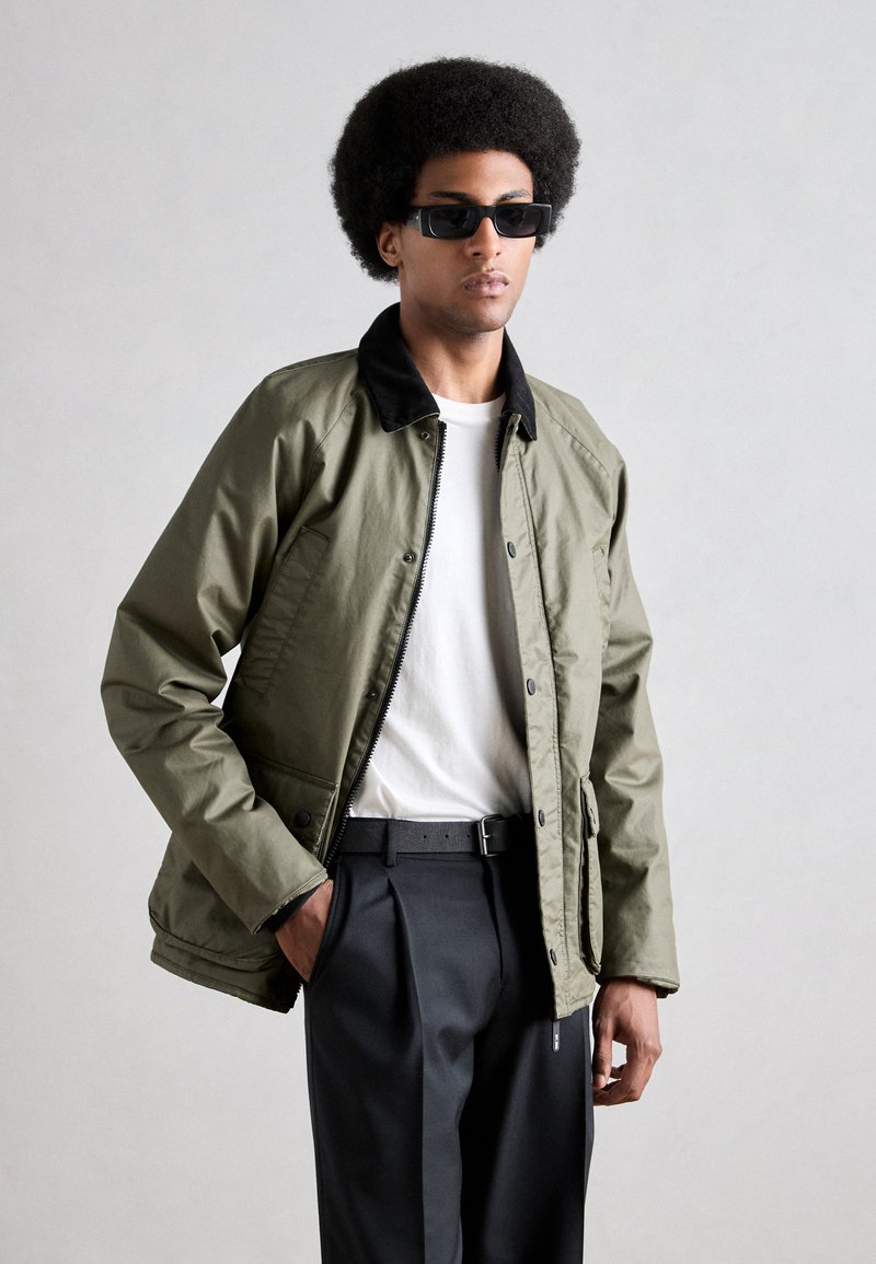 Chaqueta ligera verde oliva con cuello negro, botones en el frente y cierre de cremallera, combinada con una camiseta blanca y pantalones negros.