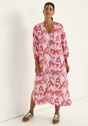 Kvinde iført en rustrød midi-kjole med knapper, dekoreret med hvide bladmønstre og trekvartærmer, kombineret med brune sneakers med hvide snørebånd.