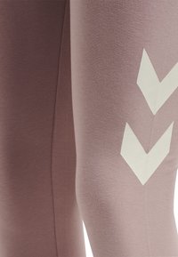 Legíny ve vybledlé mauve barvě, vyrobené z měkké, elastické tkaniny. Obsahují bílé chevron vzory po stranách a hladké švy.